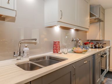 A kitchen counter with a dual sink, faucet, soap dispenser, red container, glasses, kettle, bread, pots, and stove at Maple Cottage in Keswick