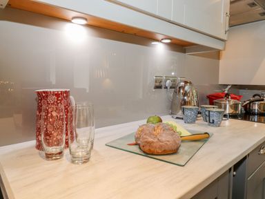 A kitchen counter with two glasses a red patterned jug bread cheese an apple green grapes a kettle three mugs and pots at Maple Cottage in Keswick