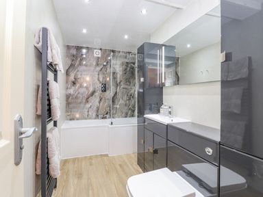 A bathroom with a bathtub shower combination a black towel rack with towels and a gray vanity unit with sink and mirror at Maple Cottage in Keswick