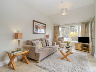 A living room with a beige sofa and armchair a glass coffee table with flowers and a wooden TV stand with a television at Garden Cottage in Keswick