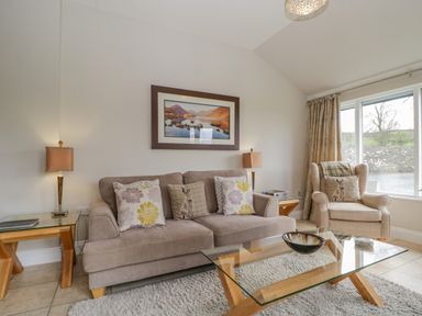 A living room with a sofa, armchair, glass coffee table, side tables, and a framed picture at Garden Cottage in Keswick