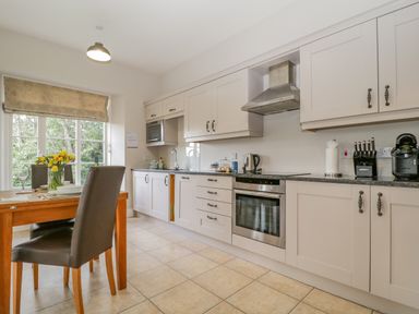 A kitchen with white cabinets a wooden dining table and chairs and a window with a blind at Garden Cottage in Keswick