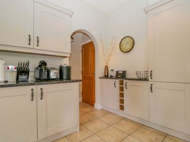 A kitchen with beige cabinets a clock on the wall appliances on the counter and a wooden door through an archway at Garden Cottage in Keswick
