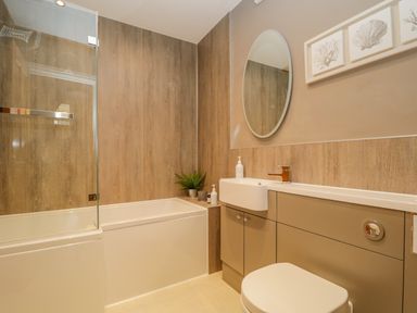 A bathroom with a bathtub shower combination a sink with cabinets a toilet a round wall mirror and framed prints on the wall at Garden Cottage in Keswick