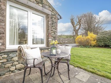 A stone patio with two metal chairs and a round metal table with cushions and a lantern next to a stone building with a window and a garden with grass and shrubs at Garden Cottage in Keswick