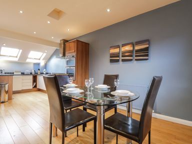 A dining area with glass table and four black chairs set with plates and glasses next to a kitchen with wooden cabinets and skylight windows at Wrenville in Keswick