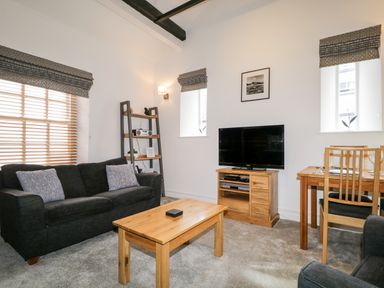 A living room with a black sofa, wooden coffee table, TV on wooden stand, shelving unit, and dining table at The Old Hayloft in Keswick