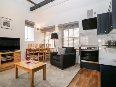 A living room with a wooden coffee table a black armchair a dining table with chairs a television and a kitchen area at The Old Hayloft in Keswick
