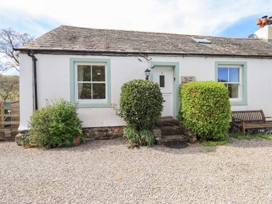 The front view of a white cottage with two windows a white door bushes and a wooden bench at Mell View Cottage in Ullswater