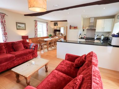 A living room with red sofas a wooden coffee table and wooden dining table near a kitchen with white cabinets at Mell View Cottage in Ullswater
