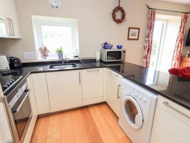 A kitchen with black countertops cream cabinets a washing machine a microwave a sink and a window at Mell View Cottage in Ullswater
