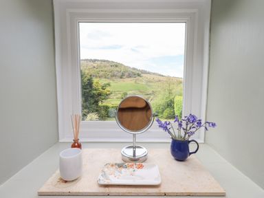 A window sill with a small mirror, blue mug with flowers, a cup, reed diffuser, and a floral plate with a countryside view at Mell View Cottage in Ullswater