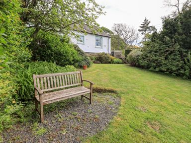 A wooden bench on gravel in a garden with grass shrubs and a white house in the background at Mell View Cottage Ullswater