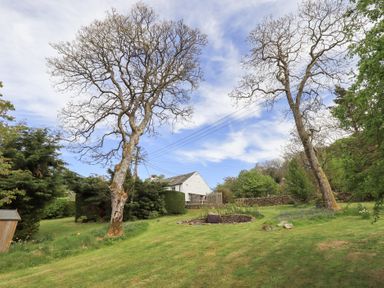A grassy garden area with trees a circular stone feature and a white house in the background at Mell View Cottage in Ullswater