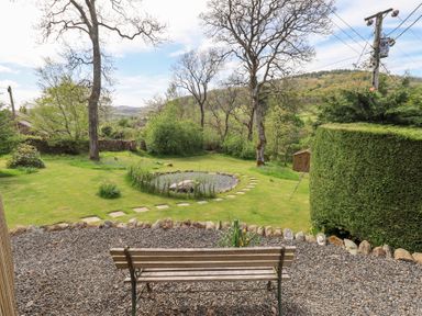 A garden with a wooden bench on gravel and a circular stone path in the grass at Mell View Cottage in Ullswater