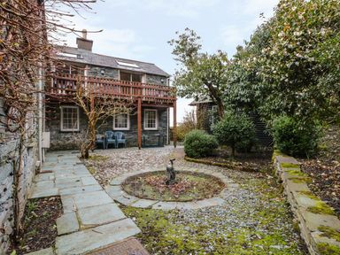 An outdoor area with a stone pathway, trees, bushes, a small fountain, and a stone building with a wooden balcony and plastic chairs at Coombe Cottage in Borrowdale