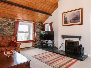 A living room with a striped sofa a wooden coffee table a flat screen tv near a fireplace with tools and a landscape painting on the wall at Coombe Cottage in Borrowdale