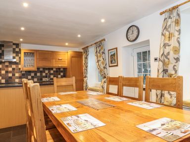 A dining area with a wooden table and chairs set with placemats next to a kitchen with wooden cabinets and tiled backsplash at Coombe Cottage in Borrowdale