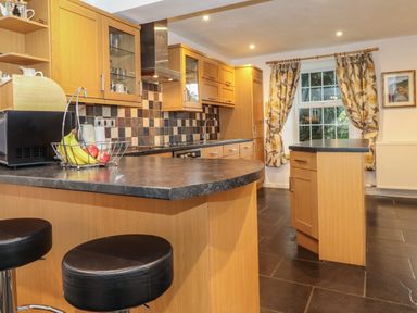A kitchen with wooden cabinets black countertops bar stools and a window with floral curtains at Coombe Cottage in Borrowdale