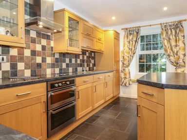 A kitchen with wooden cabinets a tiled backsplash a double oven a sink and a window with floral curtains at Coombe Cottage in Borrowdale