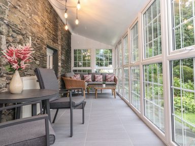 A glass sunroom with a stone wall two grey cushioned chairs a round table with flowers and a wicker sofa with cushions at Coombe Cottage in Borrowdale