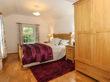 A bedroom with a wooden bed frame bedside tables lamps a wardrobe a window with floral curtains and a purple rug on a wooden floor at Coombe Cottage in Borrowdale