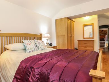 A bedroom with a wooden bed with floral pillows and a purple blanket a wooden wardrobe a chest of drawers with a mirror and a bedside table with a lamp at Coombe Cottage in Borrowdale