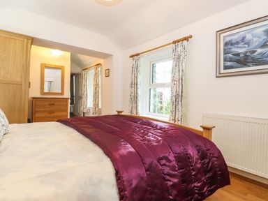 A bedroom with a bed covered with a purple blanket a wooden chest of drawers with a mirror and a window with floral curtains at Coombe Cottage in Borrowdale