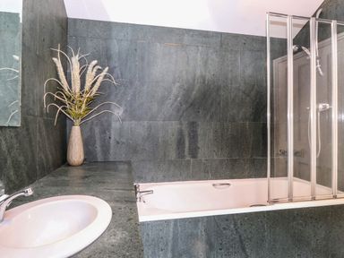 A bathroom with a sink a vase with dried plants a bathtub with a glass shower screen and dark tiled walls at Coombe Cottage in Borrowdale