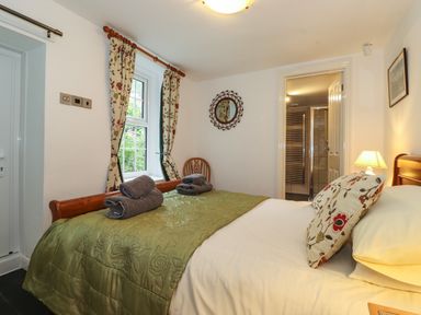 A bedroom with a bed with green cover and towels on it a wooden chair floral curtains and a door leading to a bathroom at Coombe Cottage in Borrowdale