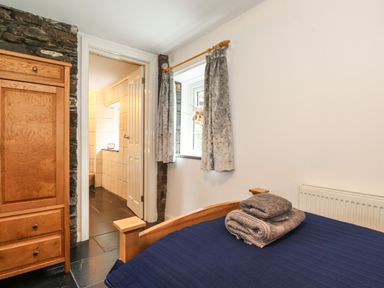 A bedroom with a wooden bed with folded towels and a wooden wardrobe near a doorway and a window with curtains at Coombe Cottage in Borrowdale