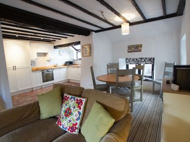 A kitchen with white cabinets and wooden countertops next to a dining area with a wooden round table and four chairs and a brown sofa with green and patterned cushions at Laurel Cottage in Spark Bridge