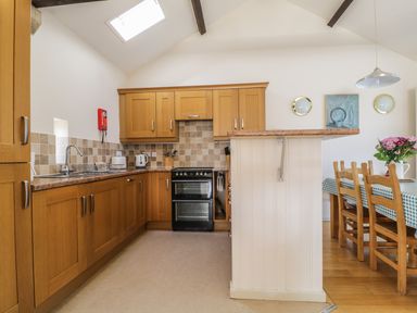A kitchen with wooden cabinets and a black oven next to a dining area with wooden chairs and a table with a checkered tablecloth and flowers at Skiddaw in Cockermouth
