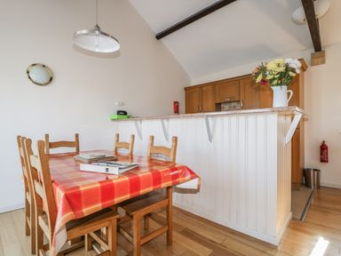 A dining area with a wooden table covered with a red checkered tablecloth and six wooden chairs next to a kitchen counter with a flower vase on top at Esthers Barn in Cockermouth