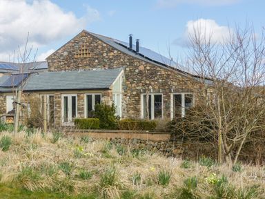 A stone house with multiple windows and solar panels on the roof surrounded by dry grass and leafless trees at Beckside in Cockermouth