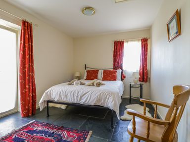 A bedroom with a bed with white linens and red pillows and curtains a wooden chair a bedside table with a lamp and a red patterned rug on the floor at Buttermere in Cockermouth