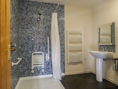 A bathroom with a walk-in shower with blue mosaic tiles a white foldable shower seat a white towel radiator a pedestal sink a wall mirror and a metal bin at Buttermere in Cockermouth
