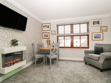 A dining area with a round table and four chairs next to a window and an armchair near a wall-mounted television above a fireplace at Andra Ghyll in Keswick