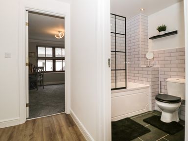 A hallway with wooden floor leading to a bathroom with a toilet black seat and a bathtub with a black framed glass screen at Andra Ghyll in Keswick