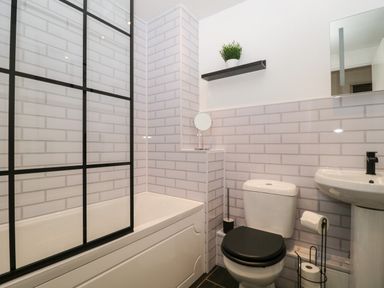 A bathroom with a bathtub and shower screen a toilet with black seat cover a sink and a black shelf with a plant at Andra Ghyll in Keswick
