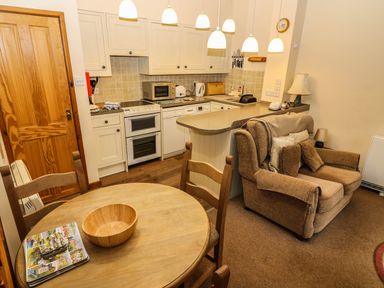 A kitchen with white cabinets a stove microwave and kettle next to a seating area with a brown armchair and a wooden dining table with chairs at Juniper Cottage in Ambleside