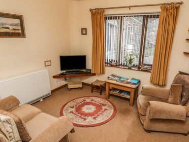 A living room with two armchairs a round rug a small wooden table with books a wall-mounted television and a window with curtains at Juniper Cottage in Ambleside