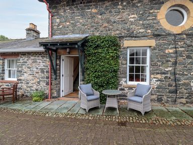 Exterior stone wall of a building with an open white door two gray cushioned chairs and a small round table at The Hayloft in Thirlmere