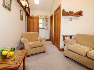 A room with two beige armchairs a wooden side table with a bowl of fruit wooden doors and a window with floral curtains at The Hayloft in Thirlmere