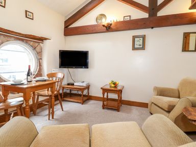 A sitting room with wooden table and chairs a TV on a stand and beige armchairs at The Hayloft in Thirlmere