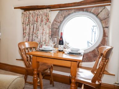 A dining area with a wooden table set for two near a round window with floral curtains at The Hayloft in Thirlmere