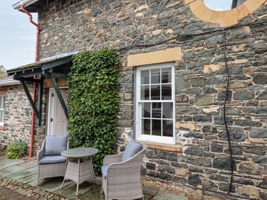 A stone exterior wall with a window and door and a small patio table with two chairs at The Hayloft in Thirlmere