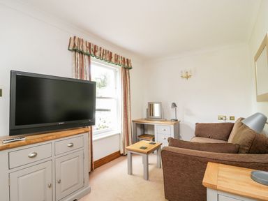 A sitting room with a brown sofa a black TV on a grey cabinet a small wooden table and a window with curtains at Thirlmere Suite in Thirlmere
