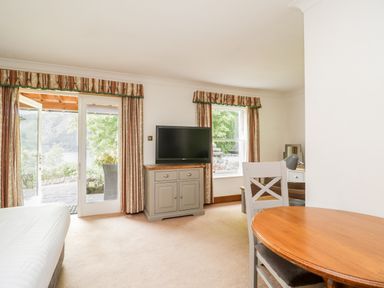 A room with a bed, wooden table with chairs, a TV on a cabinet, windows with curtains, and a door opening to a patio at Thirlmere Suite in Thirlmere