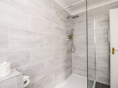 A bathroom shower area with grey tiled walls a rainfall showerhead a handheld shower a glass shower screen and a toilet paper holder at Thirlmere Suite in Thirlmere